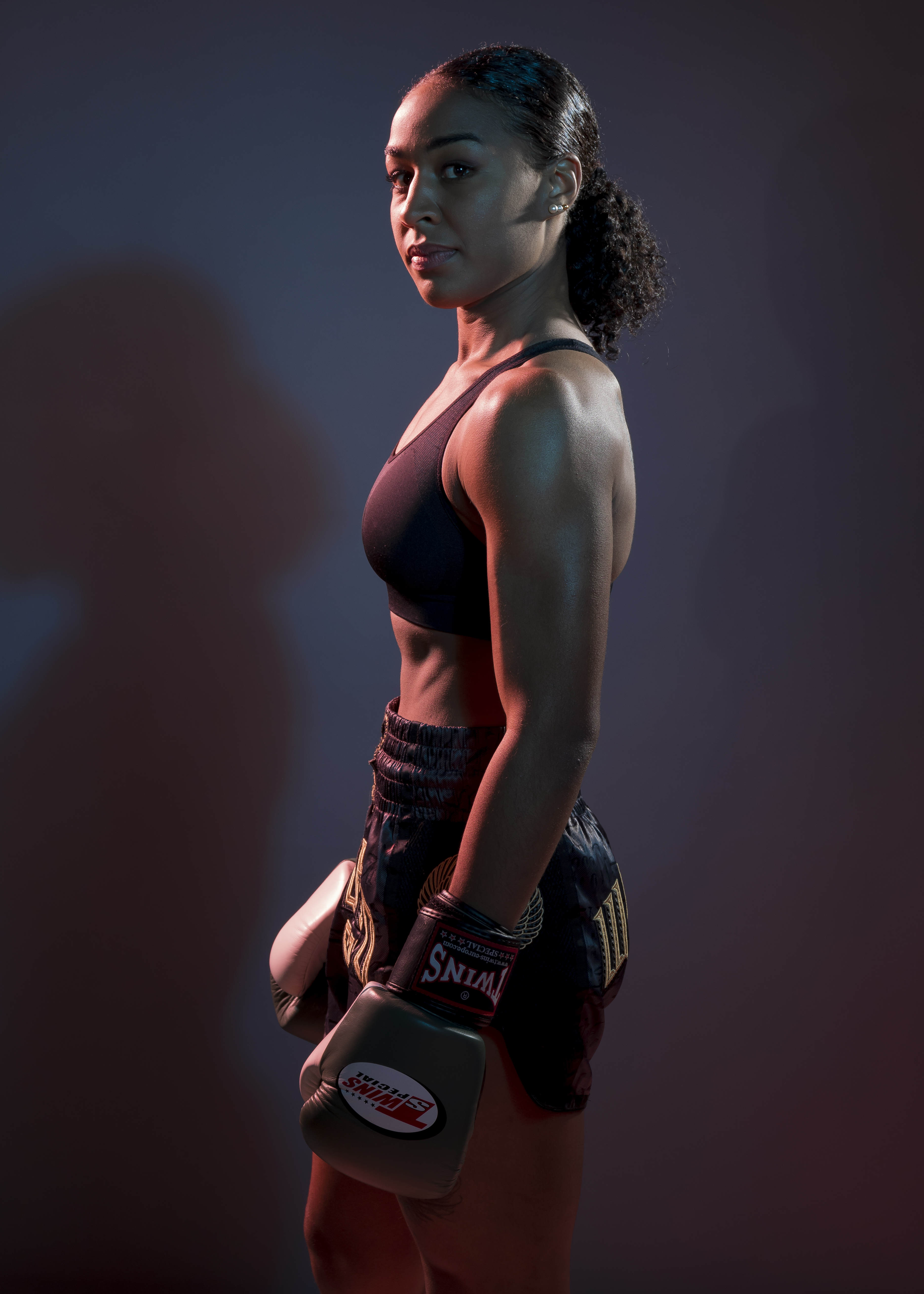 Femme athlétique en tenue de kick-boxing, gantée, regard confiant, sous un éclairage contrasté.
